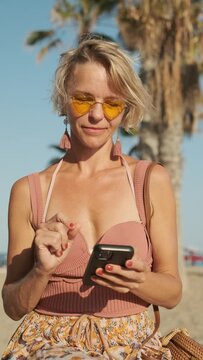 Woman with sunglasses using her mobile phone while sitting outdoors at the beach on a sunny day. Summer and technology concept.