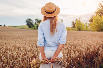 Back view of young woman enjoying summer field landscape wearing straw hat and linen shirt holding...