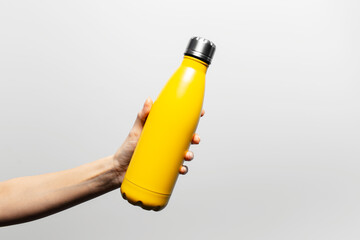 Close-up of female hand holding metal reusable thermo water bottle of yellow color on grey background.