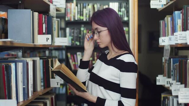 Female student with dark hair adjusts her glasses and reads a book, standing in the library, between the shelves and bookcases