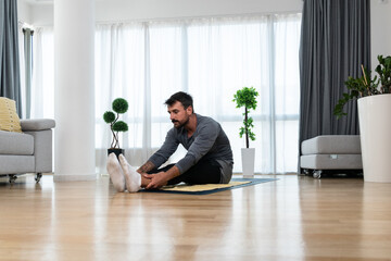 Fototapeta Happy attractive young business man in active sport wear sitting on floor stretching muscles at his apartment after or before the work, training yoga class. Home workout as stress and pain relief. obraz