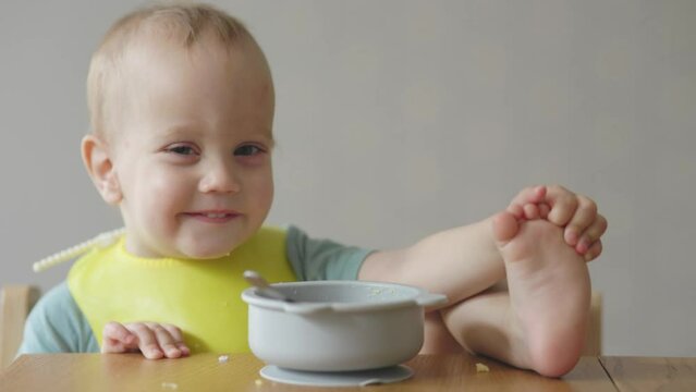 naughty baby child putting foot leg on table while eating having lunch in kitchen. happy smiling toddler kid boy caucasian playing with body near plate full of meal during dinner time. table manners