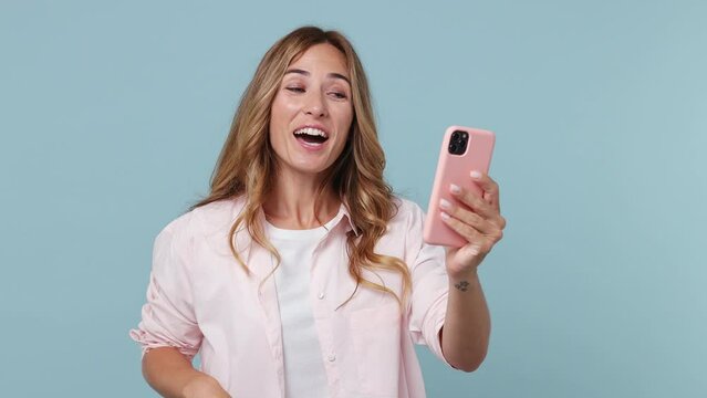 Young fun woman 30s wear pink shirt get video call using mobile cell phone doing selfie talk conducting pleasant conversation greet with hand isolated on plain pastel light blue cyan background studio