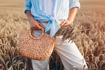 Close up of straw handbag filled with wheat. Woman holding summer purse with bundle of wheat in field at sunset - obrazy, fototapety, plakaty