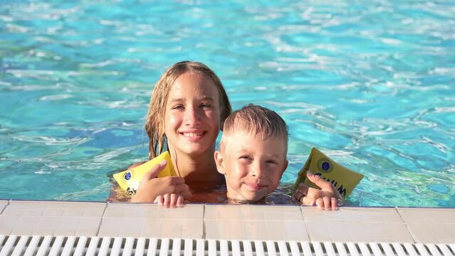 Close up of happy children swimming in pool. Brother and sister. Young beautiful girl and little boy laugh and smile broadly. Boy in inflatable armbands is holding onto side of pool. Look into camera.