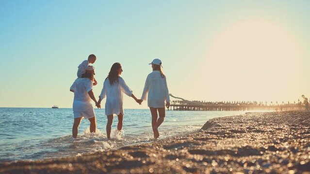 Happy family together walk holding hands on water on sea near sandy beach at sunshine day. Travel tourists. Parents and childrens leisure activity on summer outdoor . Baby son sit at shoulders dad.