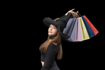 Portrait of girl in witchs hat with colorful paper shopping bags in hands. Black background. Copy...