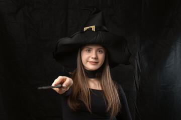 Portrait of young girl in witchs costume wears pointed hat with magic wand in her hands on black background.
