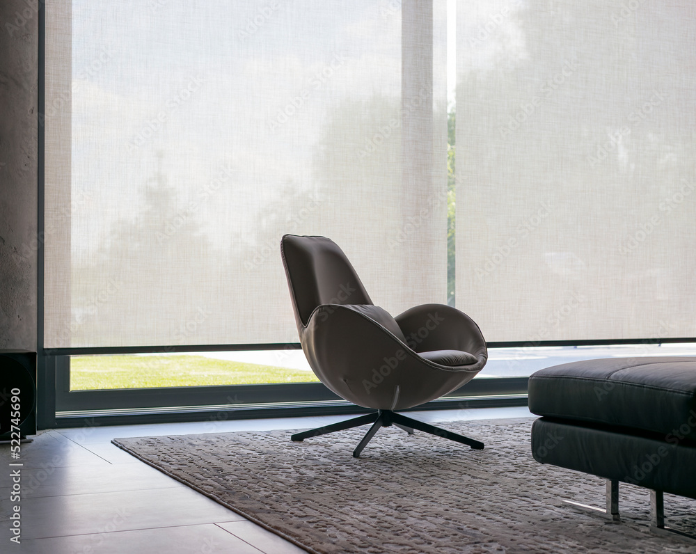 Poster Roller blinds of large sizes on the window in the interior. Automatic solar shades, fabric with linen texture. In front of a large window is a chair on a carpet. Outside is a view of the garden.