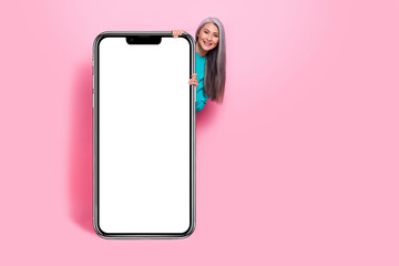Photo of funny attractive retired woman blue sweater hiding behind modern gadget empty space isolated pink color background
