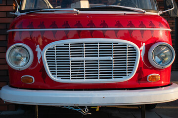 Obraz na płótnie Canvas The red body of a hippie mobile or van or bus with the pattern of a dark and white man and round headlights in the golden sunlight on the background of a brick wall. Cafe on wheels. Food truck. 
