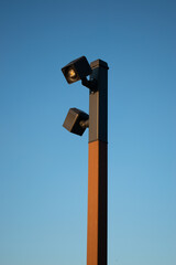 A tall insulated lantern or CCTV camera on a wooden pole or pole with an iron black or gray coating on top in golden sunlight against a clear blue sky