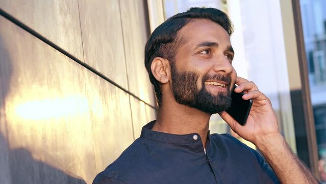 Smiling young indian business man holding smartphone talking on cell phone making call standing on urban street in modern city on sunset speaking by cellphone, laughing, having mobile conversation.