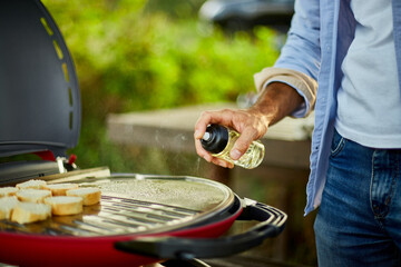 Fototapeta premium Prepare breakfast with bread, roasting bruschetta and man spraying oil on the barbecue gas grill outdoor in the backyard, Breakfast on grill, summer family picnic, food on the nature.