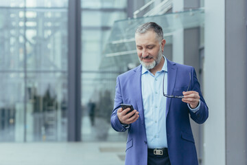 Fototapeta Successful and happy senior gray-haired businessman investor walking outside office building, man holding phone, banker using smartphone, typing message and reading news obraz