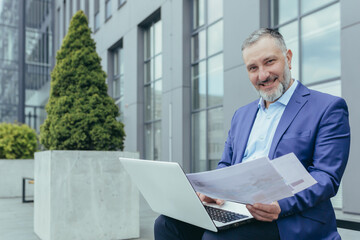 Fototapeta Portrait of successful investor behind paperwork, man outside office building sitting on bench with laptop working with documents, man in business suit smiling and looking at camera obraz