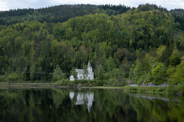 Wonderful landscapes in Norway. Vest-Agder. Beautiful scenery of whtite Gyland church reflecting in the lake. Mountains, road and trees in the background. Wooden floating platform. Selective focus