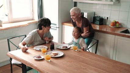 Happy mature lesbian couple talking and eating breakfast with adopted Asian daughter in the kitchen at home