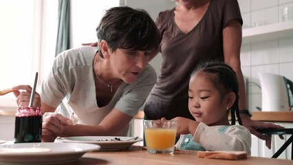 Lovely mature lesbian couple eating breakfast with adopted Asian daughter in the kitchen at home