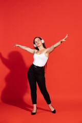 Fototapeta premium Portrait of cheerful young woman posing, listening to music in headphones isolated over red studio background