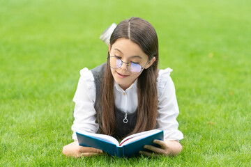 Serious schoolgirl reading book lying on grass, reading. Teen girl reading outdoors