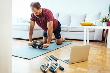 Fototapeta Mature Adult Man Exercising at Home in His Living Room. Mature man exercising at home - watching virtual exercise class on laptop. . Athletic man in sports clothes having domestic training obraz