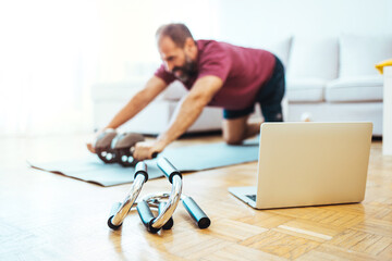 Fototapeta Man watching gym class on laptop and doing exercises at home. Positive athletic bearded middle-aged man exercising at home, using laptop, watching sport videos on Internet. obraz
