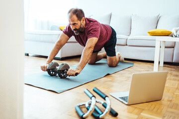 Fototapeta Positive athletic bearded middle-aged man exercising at home, using laptop, watching sport videos on Internet, having fitness class online, copy space. Healthy lifestyle, sport on self-isolation obraz
