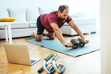 Fototapeta Senior man working out at home, watching youtube videos and learning the exercises. Mature man exercising at home - watching virtual exercise class on laptop. obraz