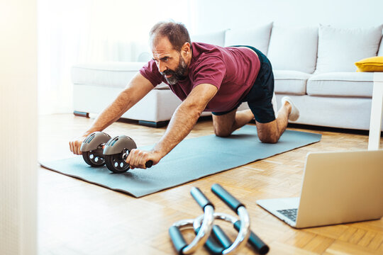 Senior Man Working Out At Home, Watching Youtube Videos And Learning The Exercises. Mature Man Exercising At Home - Watching Virtual Exercise Class On Laptop.