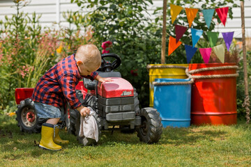 A cheerful boy dressed in a rustic style washes a tractor with a rag in the garden in the village....