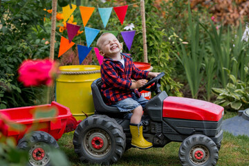 A cheerful boy dressed in a rustic style is sitting on a tractor in a garden in the village....
