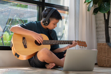 Asian boy playing guitar and watching online course on laptop while practicing for learning music...