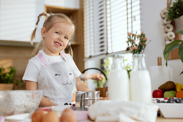 Happy preschooler girl wearing apron in kitchen helping mother cooking baking cookies muffins preparing surprise for family standing at table in beautiful modern kitchen having fun with mom smiling.
