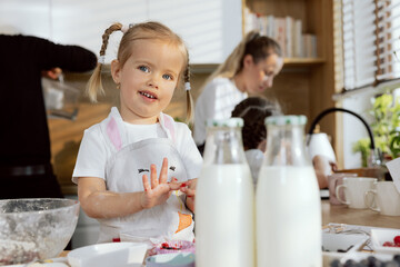 Little curious girl standing at table in kitchen collecting remnants of jam from hands kneading dough cooking baking tasty cookies with family in background for breakfast.
