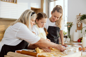 Adorable young girl wearing apron pressing cookies with cookies cutter helping granny and mother...