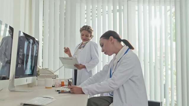Female doctor examining x ray image on computer and taking notes on clipboard as her colleague talking on phone at work in clinic