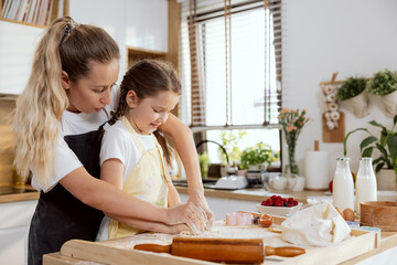 Young beautiful mother teaching little adorable daughter kneading dough preparing homemade bread...
