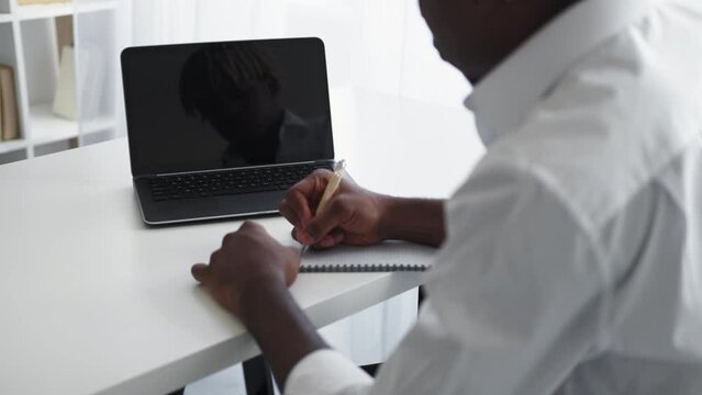 Online education. Studying man. Virtual connection. Elegant black guy sitting work desk with opened laptop dark screen writing in notebook light room interior.