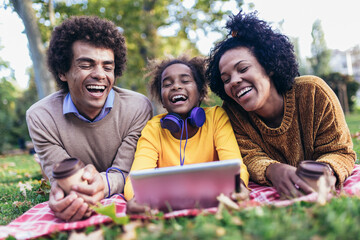 Beautiful young family lying on a picnic blanket, enjoying an autumn day in park while using digital tablet.