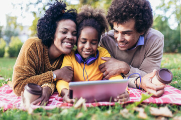Beautiful young family lying on a picnic blanket, enjoying an autumn day in park while using digital tablet.