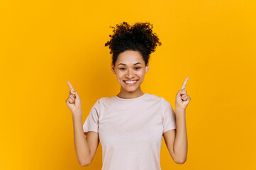 Positive friendly african american curly haired young woman, looks at the camera and points fingers up, at empty copy space, stands on isolated orange color background, smiling happily