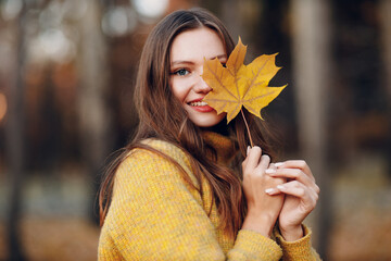 Beautiful smiling girl in warm sweater at autumn park hold in hand yellow maple foliage leaf.