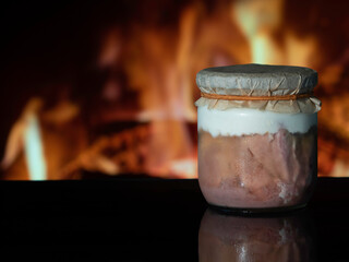 A jar of home-made canned meat with a parchment cover on a black surface in the background of a fireplace fire