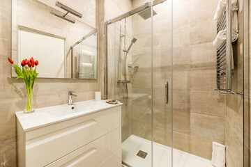 Bathroom with glass sliding shower stall, vase with artificial tulips, white dresser, marble tiles and frameless mirror