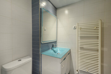 Bathroom with frameless mirror with blue edges, blue glass sink, white chest of drawers and white towel radiator