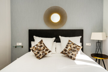 Bedroom with headboard upholstered in black fabric, cushions and pillows, decorative mirror and white lamp on round table
