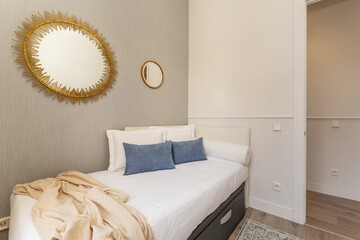 Bedroom with a single bed with gray leatherette upholstered drawers, white and blue cushions and decorative mirrors with gilt frames