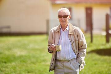 Serious elderly man 75 years old standing outdoors in front of his house with mug of hot drink in...