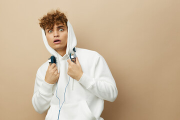 a cute, attractive man stands on a beige background in a white hoodie on his head and holds...
